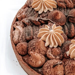 Auf dem Bild ist eine Draufsicht auf eine Schokoladen Karamell Brownie Tarte. Die Oberfläche der Tarte ist reich verziert und besteht aus dressierter Schokoladenganache, die in verschiedenen Formen aufgetragen wurde. ### Formen der dressierten Schokoladenganache: 1. **Halbkugeln:** Es gibt mehrere symmetrisch platzierte, kleine Halbkugeln aus Schokoladenganache. Sie sind gleichmäßig und haben eine glatte, runde Form. 2. **Schwungvolle Linien:** Teile der Ganache sind in geschwungenen, fast wellenartigen Mustern aufgetragen. Diese Linien sind dicht aneinander und verlaufen in einer Art horizontalen Bewegung. 3. **Stachelige Strukturen:** Einige Abschnitte zeigen Spitzen oder fast gezackte Formen, die der Ganache Textur und Dimension verleihen. 4. **Punkte und Tupfer:** Zwischen den größeren Mustern befinden sich kleine, punktartige Details aus Ganache, die wie Tropfen wirken. 5. **Cremeblüten:** Auf der Torte befinden sich mehrere große, symmetrische Blüten aus aufgeschlagener Karamell Ganache die kontrastreich zur dunklen Schokoladenganache wirken.