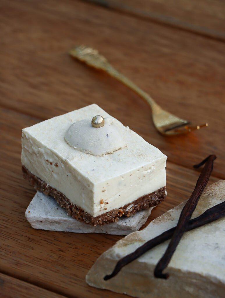 Das Bild stellt ein rechteckiges Stück von einem no Bake Vanille Cheesecake dar. Die untere Schicht ist ein crispy crunch keks-haferflocken-kokos-boden. Darüber liegt direkt die Cheesecake Schicht aus Vanille, getoppt von einer whipped vanille Ganache mit goldener Dekokugel. Daneben liegen zwei Vanilleschoten.