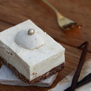 Das Bild stellt ein rechteckiges Stück von einem no Bake Vanille Cheesecake dar. Die untere Schicht ist ein crispy crunch keks-haferflocken-kokos-boden. Darüber liegt direkt die Cheesecake Schicht aus Vanille, getoppt von einer whipped vanille Ganache mit goldener Dekokugel. Daneben liegen zwei Vanilleschoten.