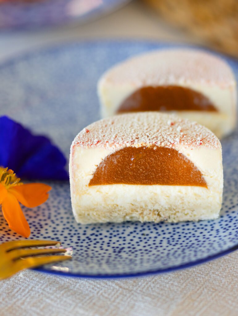 Anschnitt von einem frühlingshaften Moussetörtchen mit Jasminmousse und einem fruchtigen Aprikosenkern auf einem blauen teller und orange farbener Blüte
