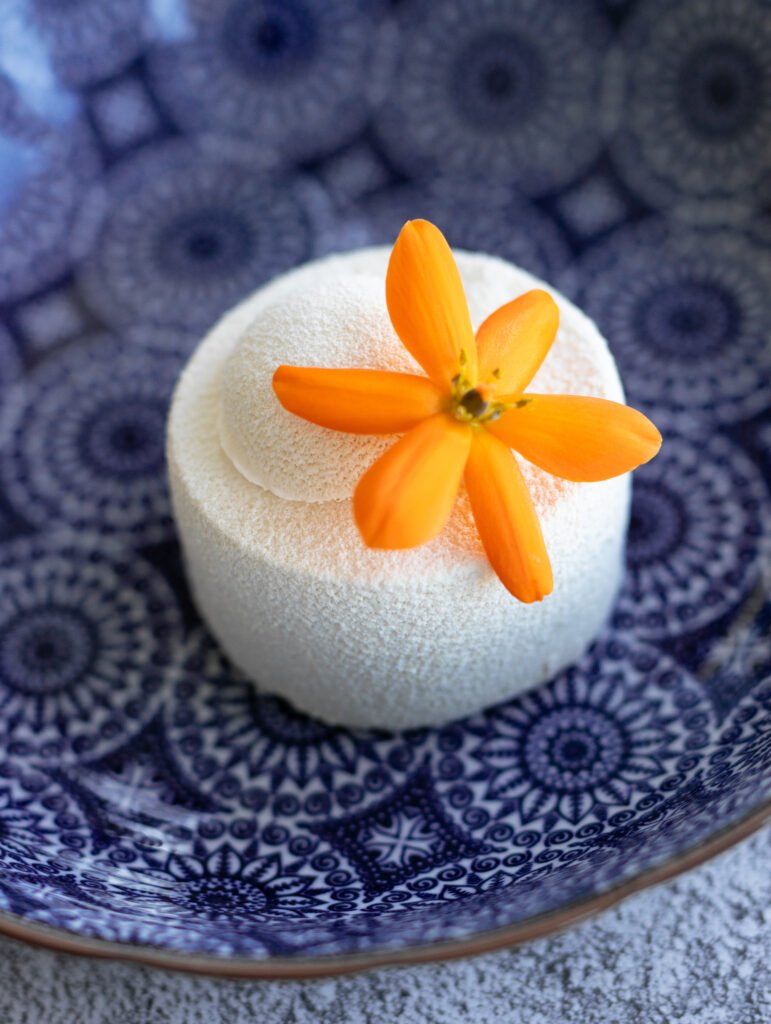 Draufsicht auf ein Moussetörtchen mit weißem Velvet Überzug aus Schokolade und Kakaobutter, mit einer orangefarbener Blüte drauf