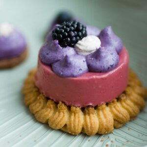 Dieses Törtchen besteht aus einer fruchtigen Waldbeermousse auf zartem Pâte Sucrée Boden, gefüllt mit einem intensiven Brownie-Inlay. Eine violette Ganache aus wilden Heidelbeeren bildet den Abschluss, gekrönt von einer Brombeere. Das Bild zeigt den Anschnitt und die raffinierten Schichten im Inneren – ein elegantes Dessert für anspruchsvolle Genießer.