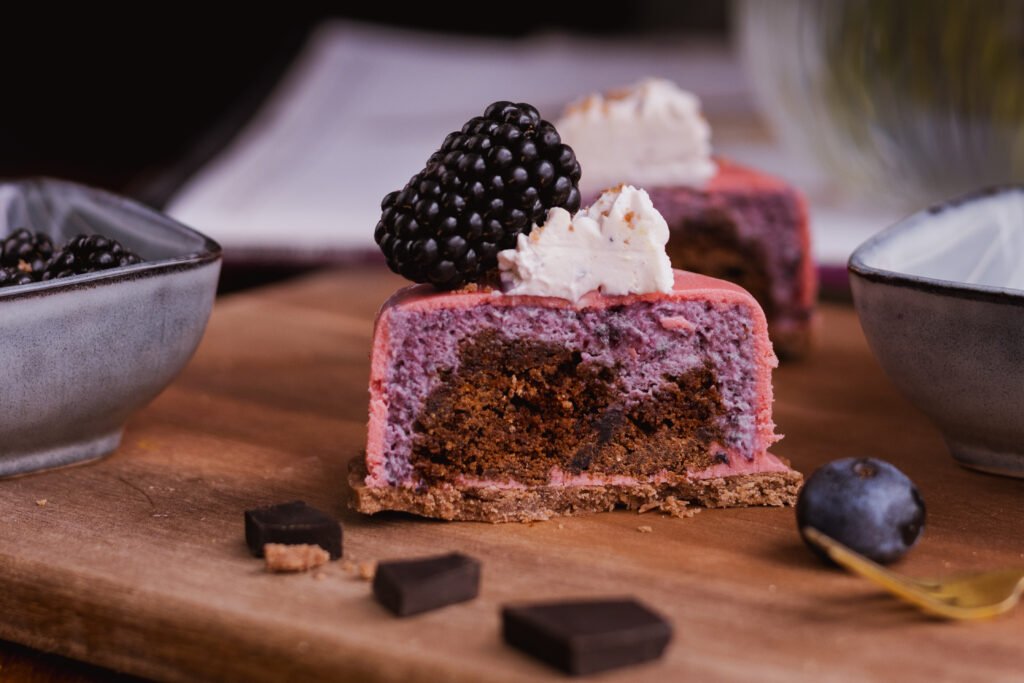 Anschnitt eines Mousse-Törtchens auf einem Pate Sucree Boden mit Brownie-Cookie-Kern, Waldbeermousse und Frischkäse-Topping, einer Brombeere und Schokostückchen auf Holzbrett